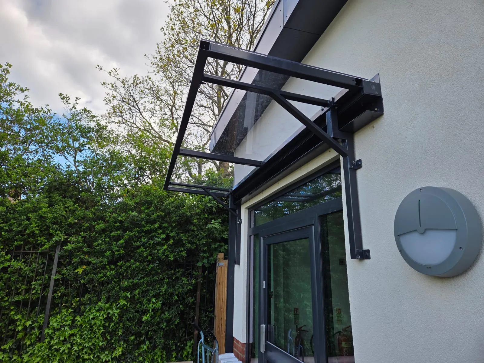 Glazed lean-to entrance canopy with black steel frame over a doorway, surrounded by mature hedging